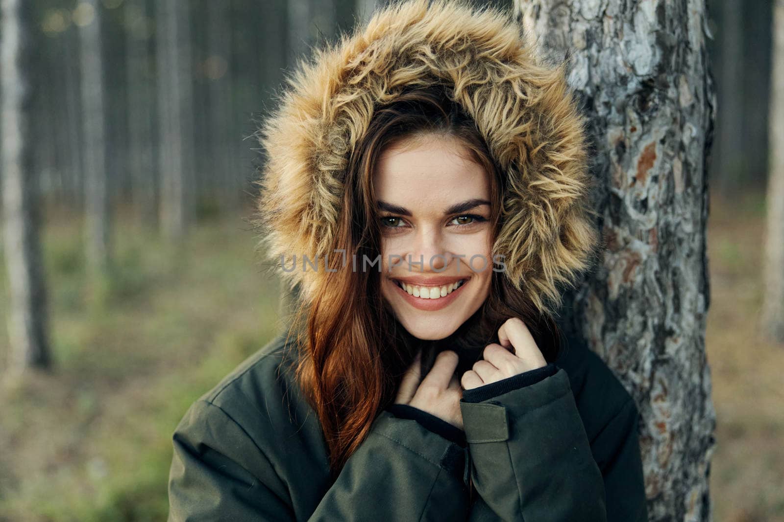 smiling woman in a jacket with a hood near a tree in nature by SHOTPRIME
