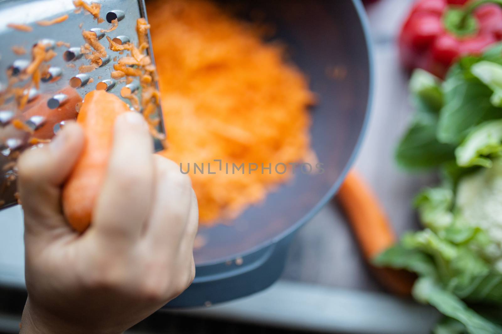 Female hands firmly grating carrot on metal grater and into bowl. Shredded carrots in bowl surrounded by vegetables. Vegan meal preparation