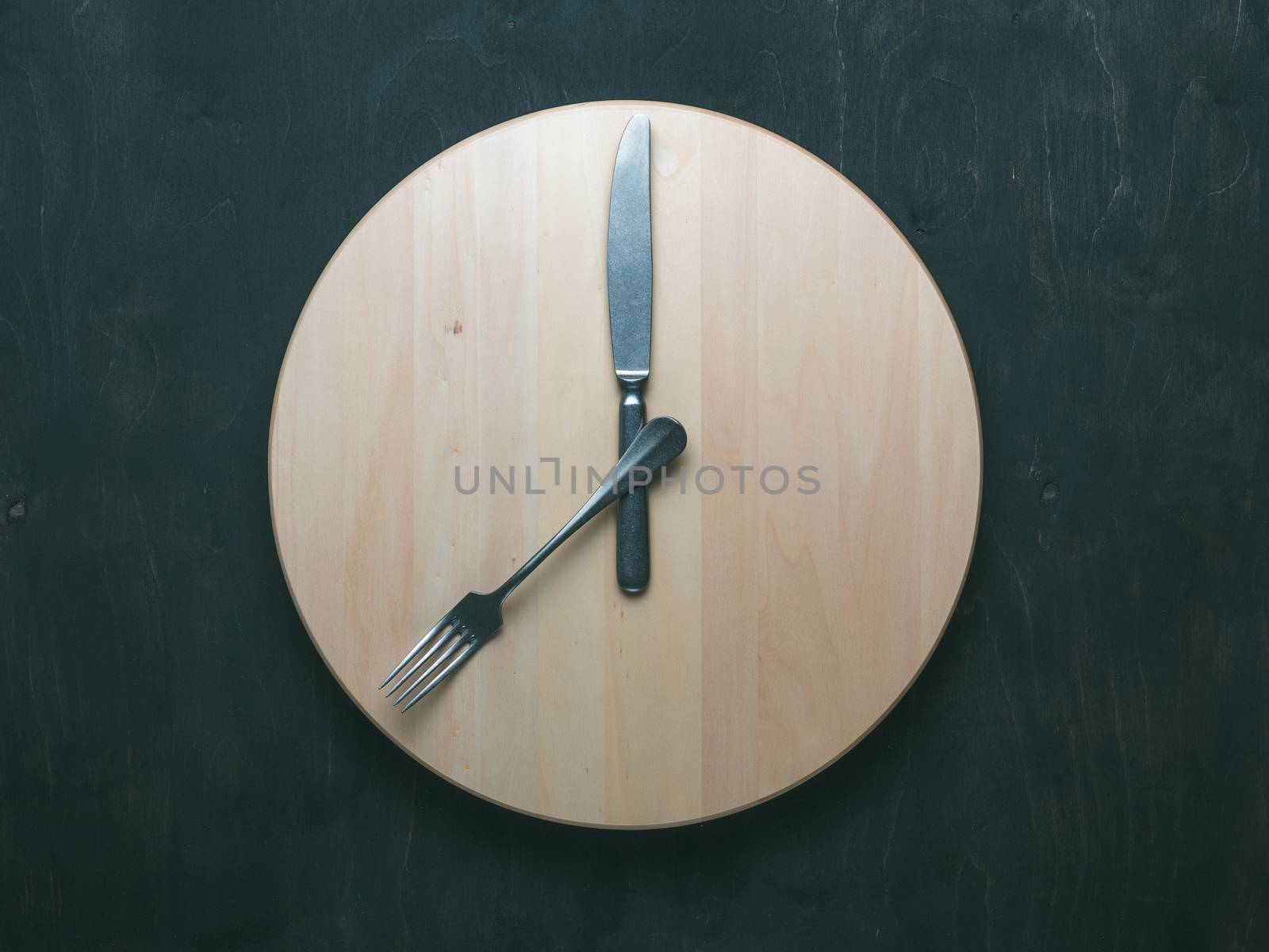 Intermittent fasting and skip breakfast concept - empty wooden round tray or trencher with cutlery as clock hands on dark background. Eight hour feeding window concept or breakfast time concept