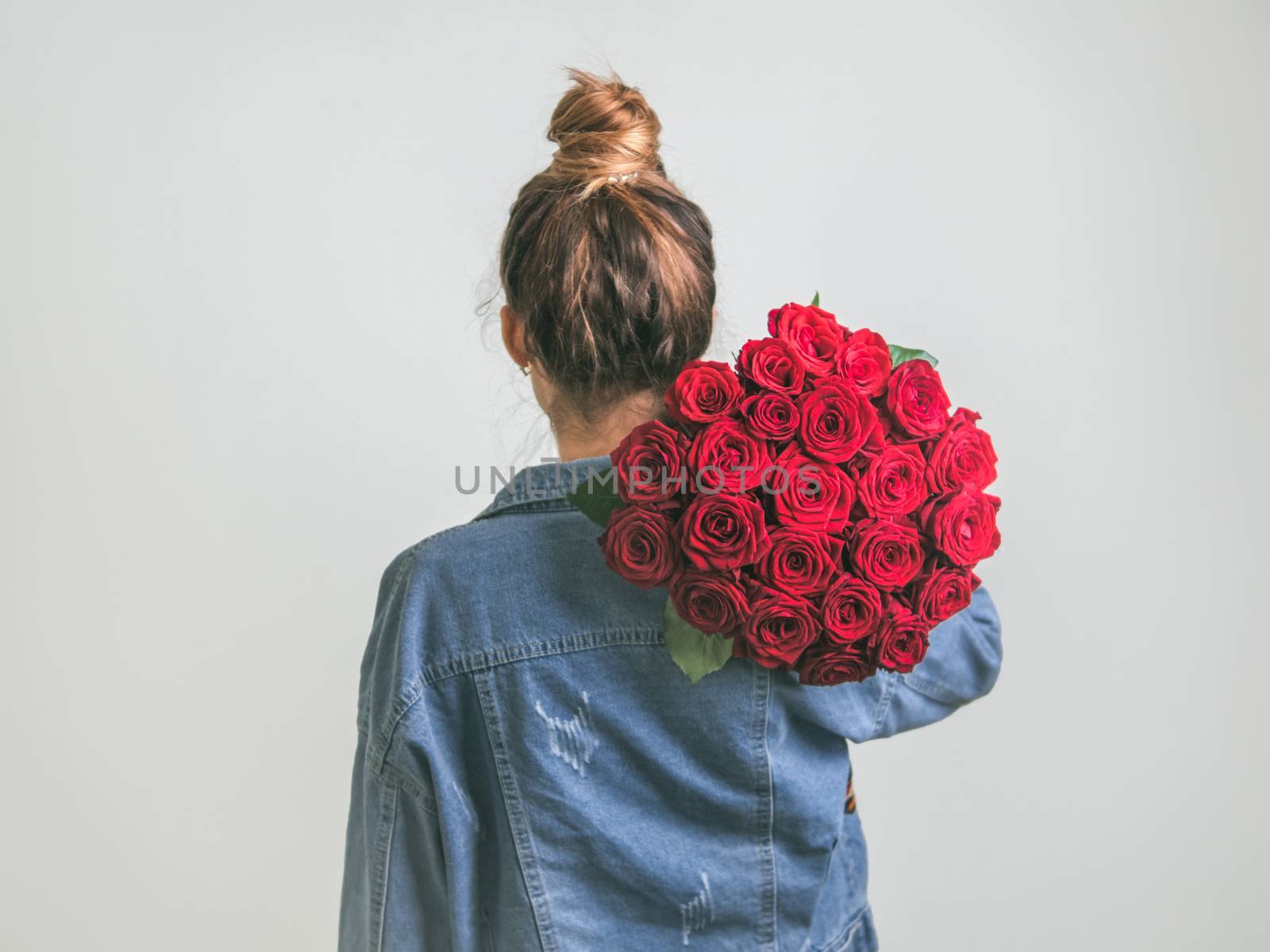 Back view of young woman in denim jacket holding bunch of red roses on shoulder. Girl with bun updo in jeans holding flowers. White background.Copy space. Visual content and idea for social media post