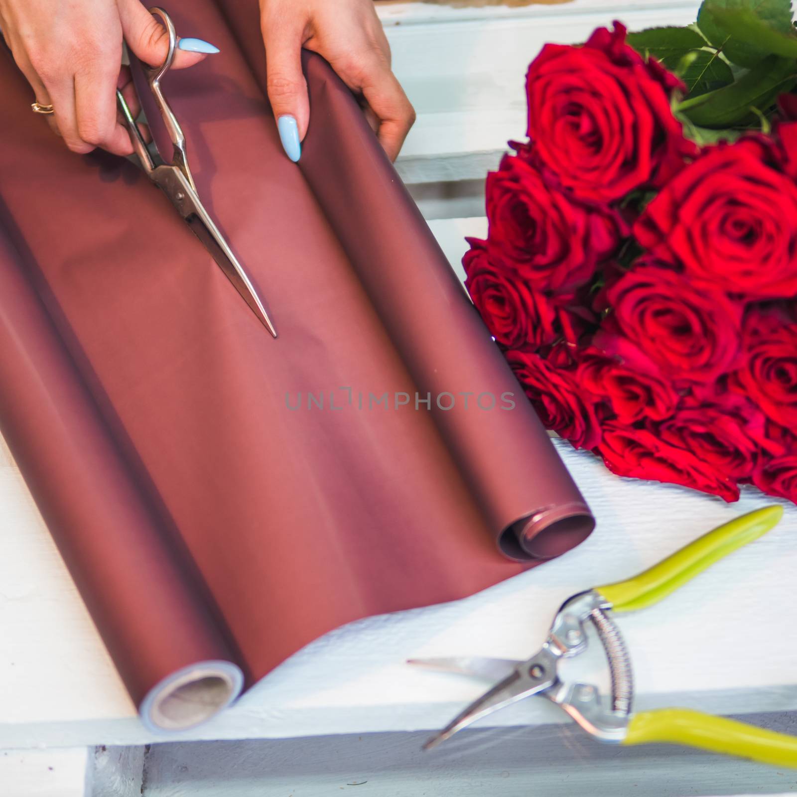 florist hands are cutting wrapping paper with scissors for making bouquet. Florist at work, making red rose bouquet in flower shop. Copy space.