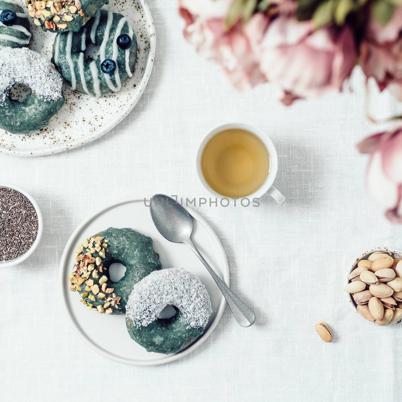 Vegan doughnuts with chia seeds topped with healthy spirulina glaze with pistachio, desiccated coconut and blueberry. Blue green spirulina donuts, herbal tea cup on table with white linen tablecloth.