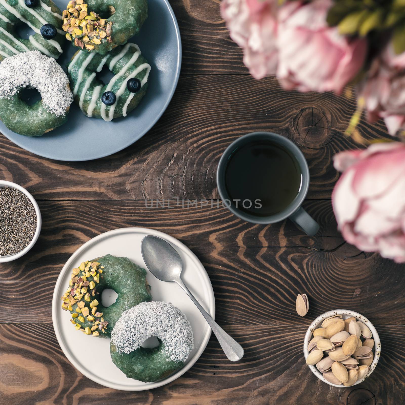 Vegan doughnuts with chia seeds topped with healthy spirulina glaze with pistachio,desiccated coconut and blueberry.Blue green spirulina donuts, herbal tea cup on brown wooden table.Top view,flat lay