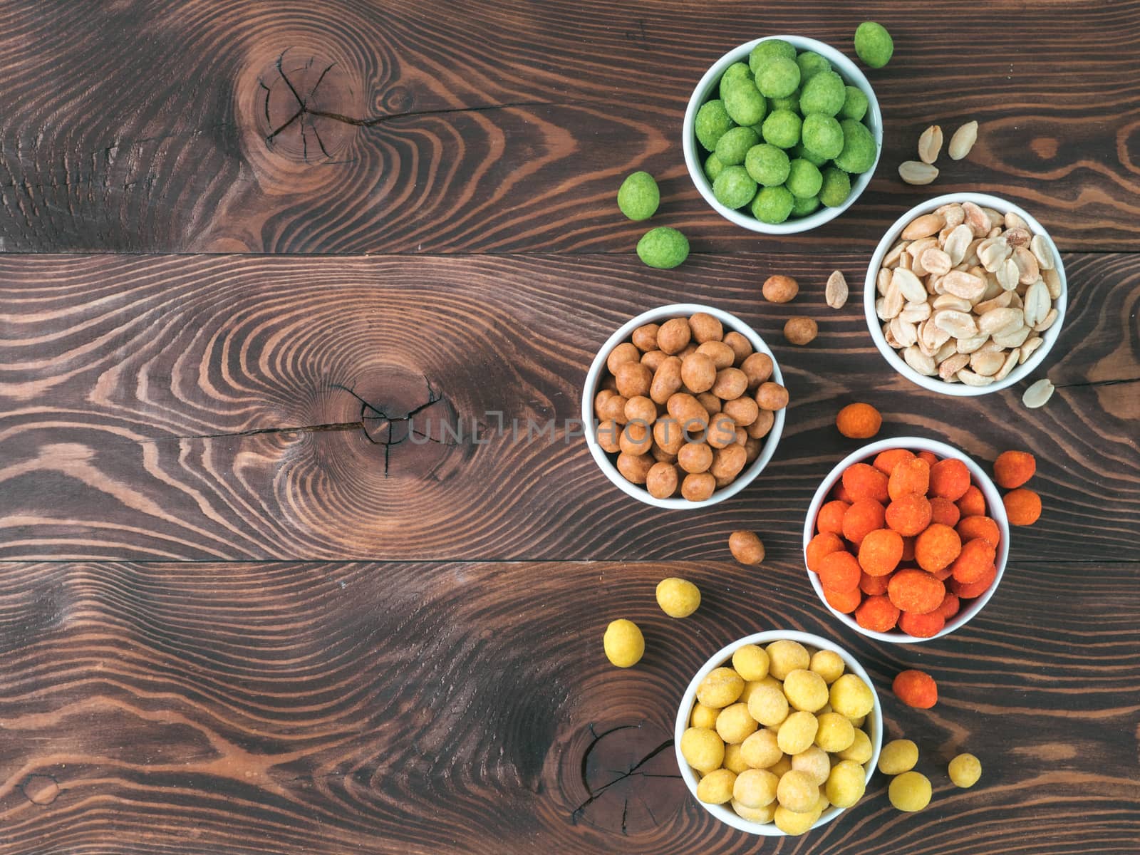 Assortment of different peanuts snack for beer, wine, party bar. Salted peanuts, peanuts in coconut glaze, green vasabi, red spicy chilli, yellow cheese glaze on brown wooden table. Copy space.