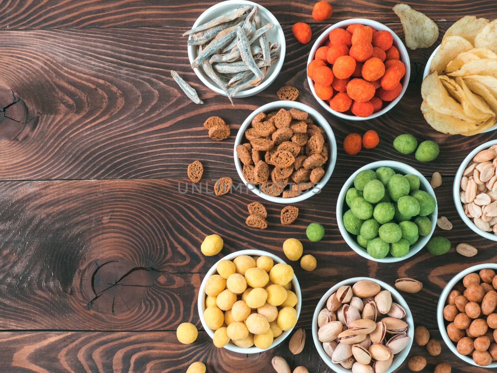 Assortment of different snack for beer, wine, party. Peanuts in coconut glaze, green vasabi, red spicy chilli, yellow cheese glaze, chips, pistachio, crackers, fish on brown wooden table. Copy space