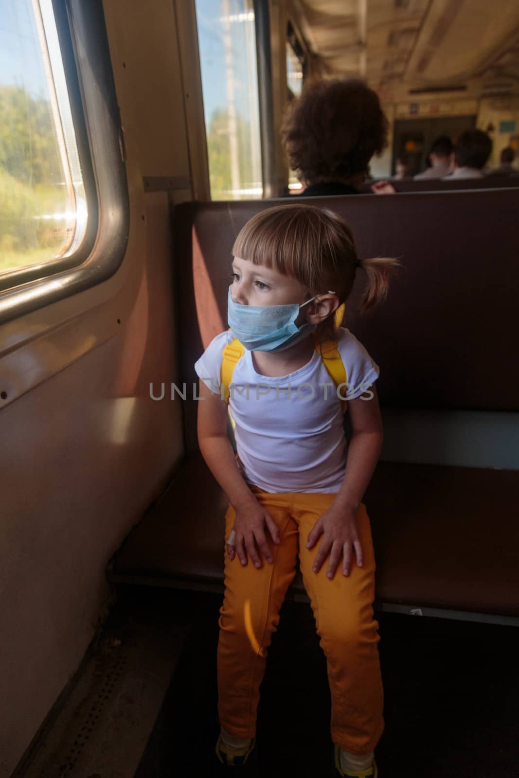 Little girl in a white T-shirt and jeans and a medical mask is sitting in the train. Keep social distancing to avoid the spread of COVID-19. Safe travel.