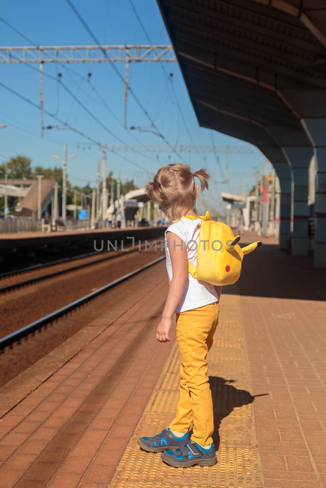 Little girl in a white T-shirt and jeans and a medical mask waiting for the train on the railway station. Keep social distancing to avoid the spread of COVID-19. Safe travel.