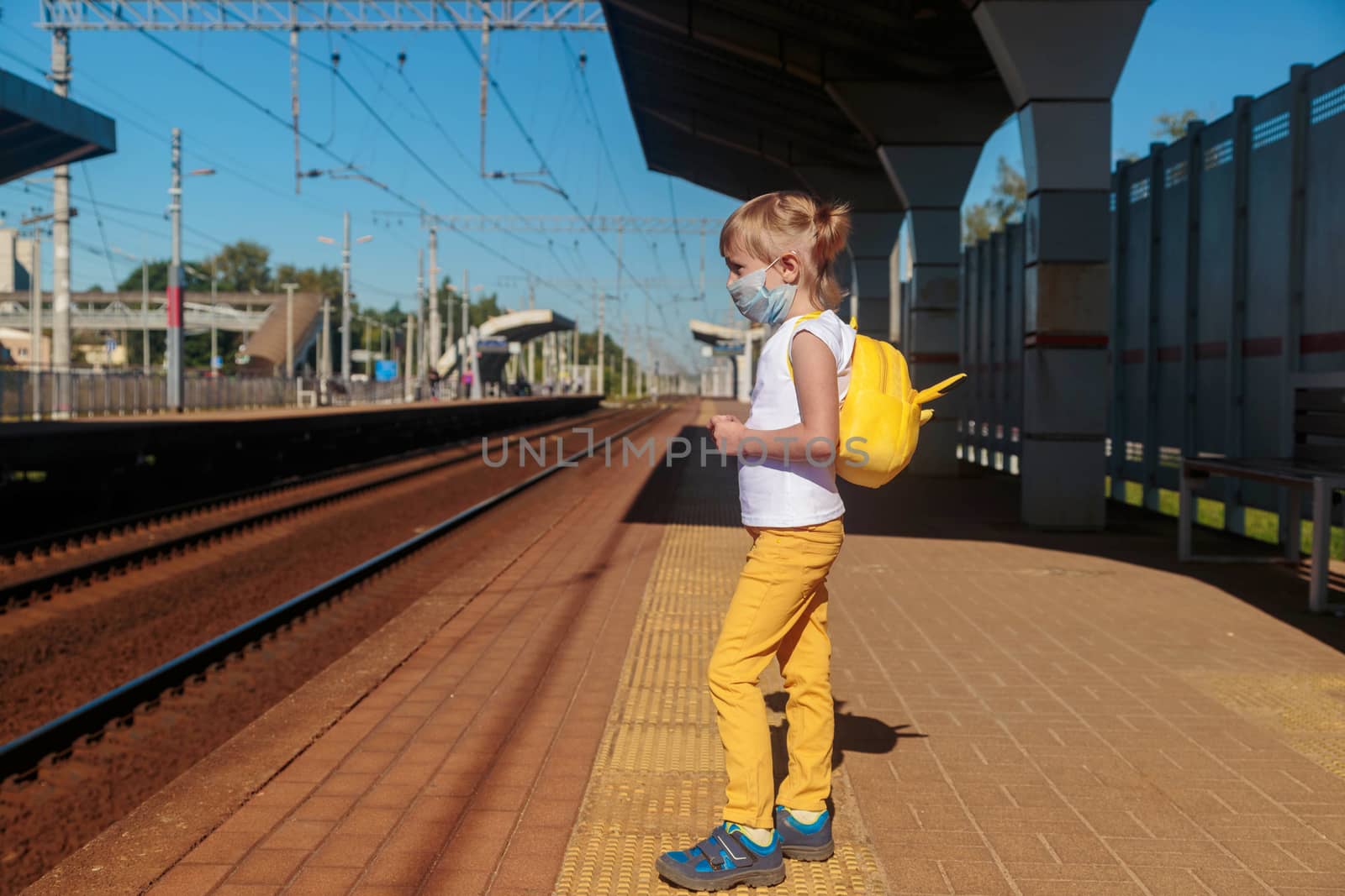 Little girl in a T-shirt and jeans and a medical mask waiting for the train by galinasharapova