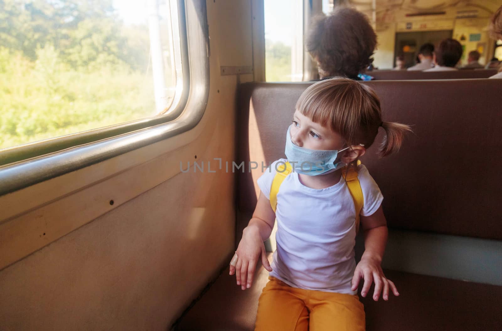 Little girl in a white T-shirt and jeans and a medical mask is sitting in the train. Keep social distancing to avoid the spread of COVID-19. Safe travel.