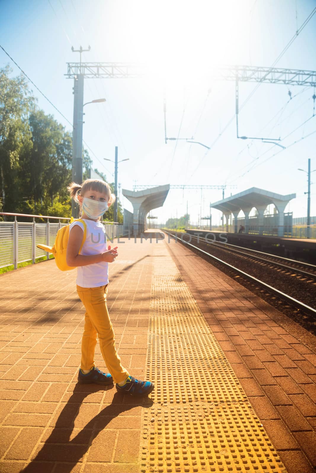 Little girl in a white T-shirt and jeans and a medical mask waiting for the train on the railway station. Keep social distancing to avoid the spread of COVID-19. Safe travel.