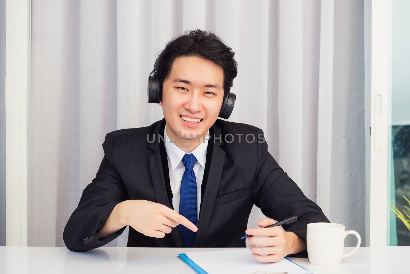 Work from home, Asian young businessman wear suit video conference call or facetime he smiling sitting on desk wearing headphones and raise his hand point finger to a paper job he looking to camera