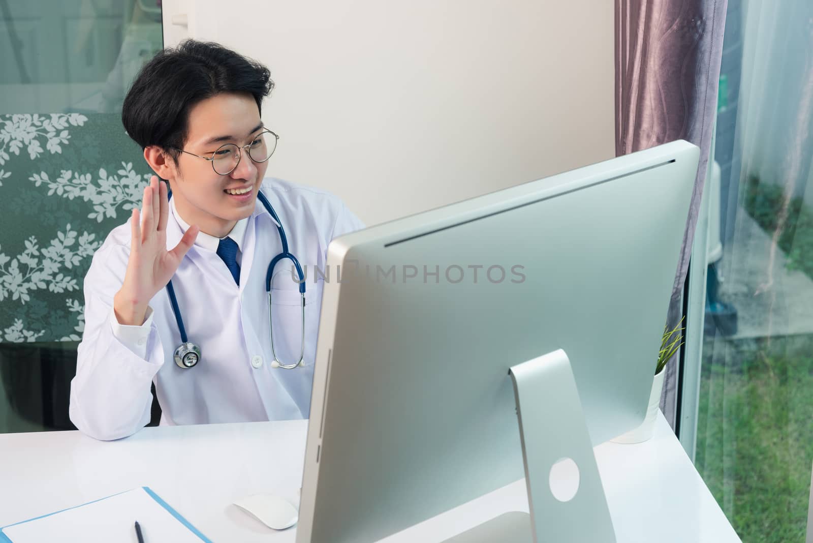 Asian young handsome doctor man wearing a doctor's dress and stethoscope video conference call or facetime raise hand say hello to patient he smiling on desk at hospital office, Health medical care