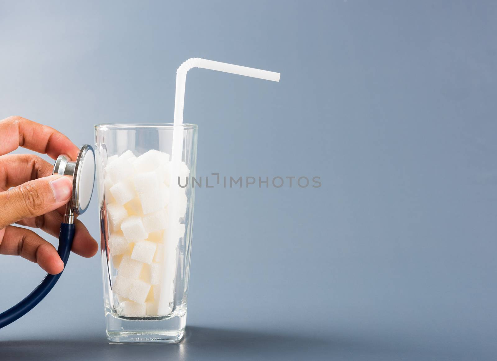 Hand of doctor hold stethoscope check on glass full of white sugar cube sweet food ingredient, isolated on gray background, health high blood risk of diabetes and calorie intake unhealthy drink