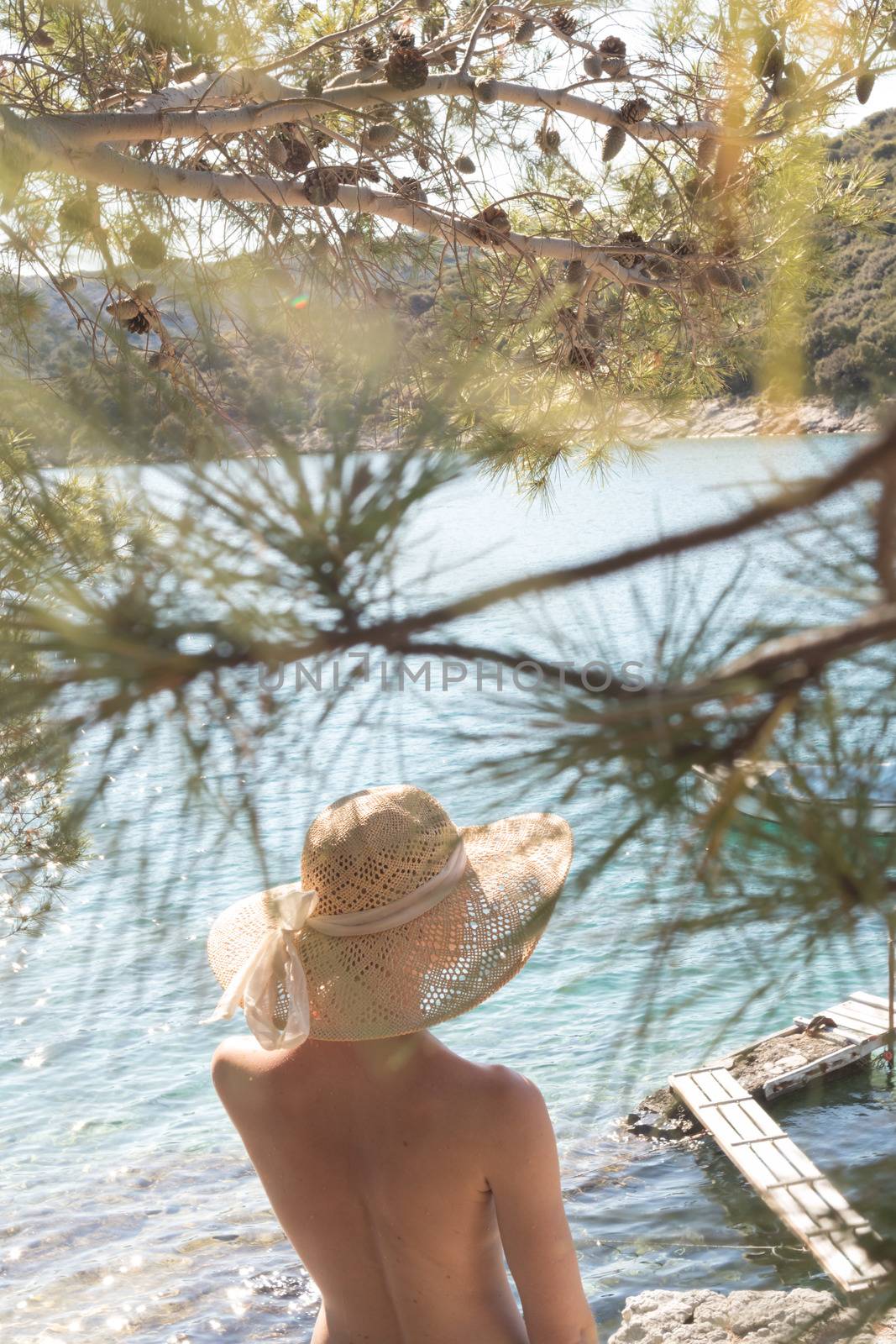 Rear view of topless beautiful woman wearing nothing but straw sun hat realaxing on wild coast of Adriatic sea on a beach in shade of pine tree. Relaxed healthy lifestyle concept.