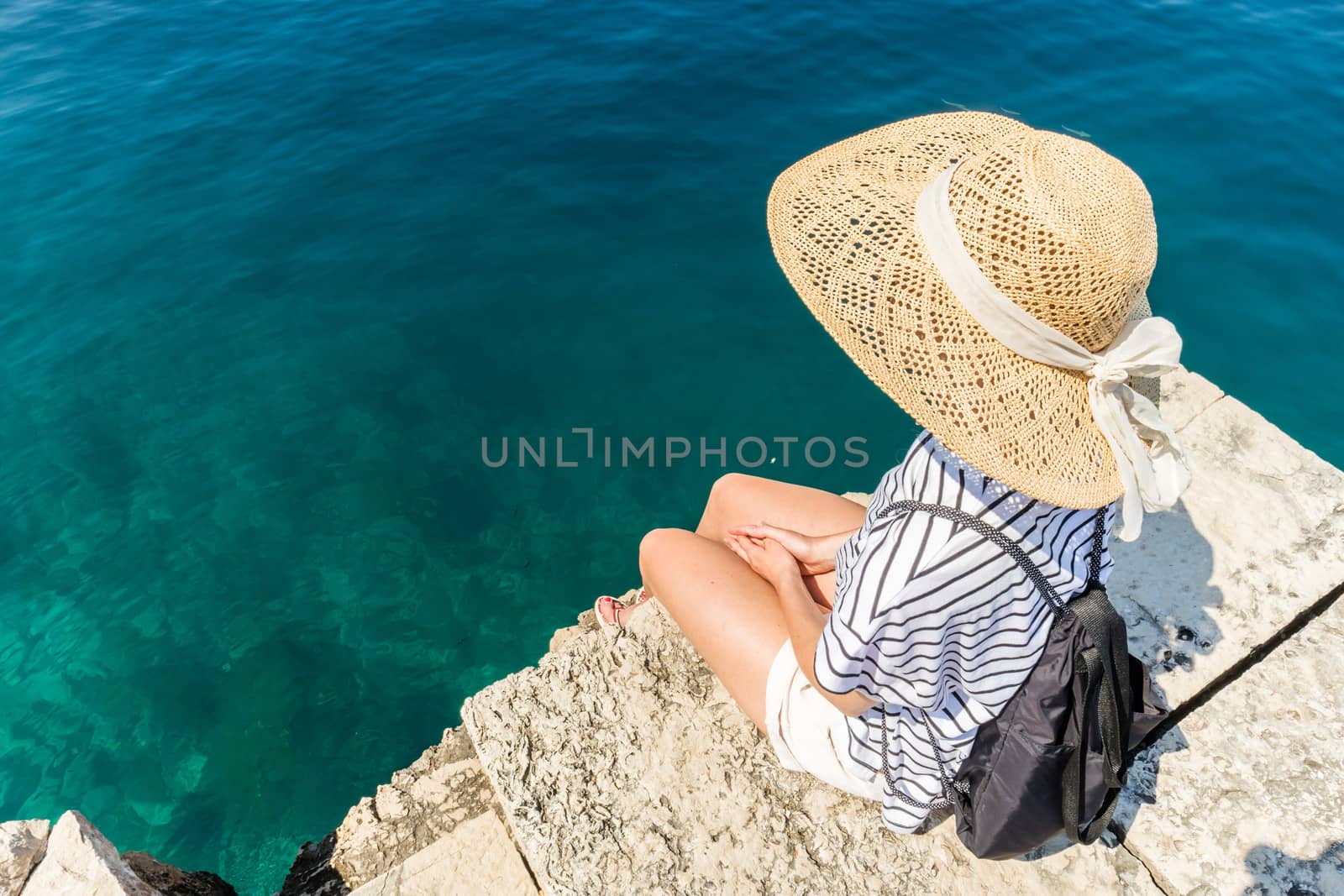 Woman traveler wearing straw summer hat and backpack, standing at edge of the rocky cliff looking at big blue sea and islands in on the horizon by kasto