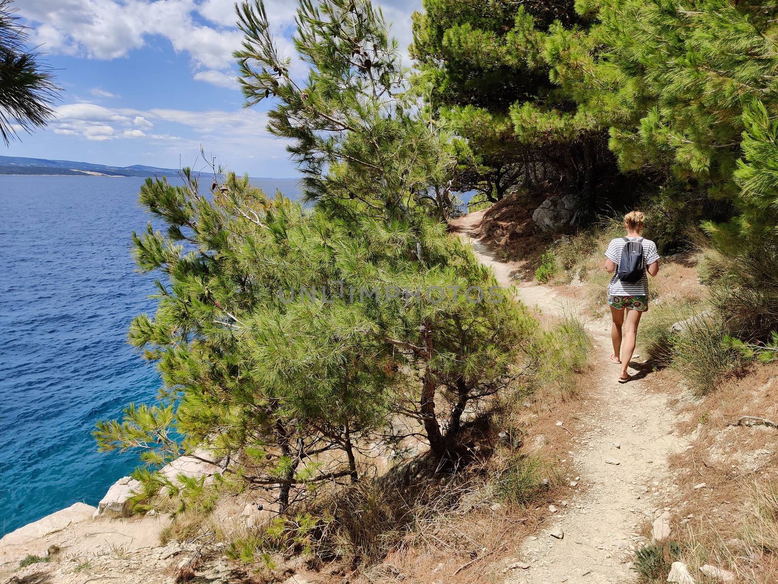 Young active feamle tourist wearing small backpack walking on coastal path among pine trees looking for remote cove to swim alone in peace on seaside in Croatia. Travel and adventure concept.