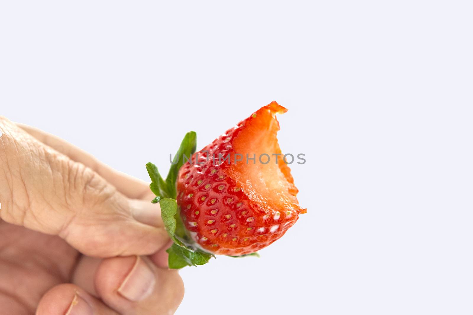 Bitten off red strawberry with green leaves in hand against white background
