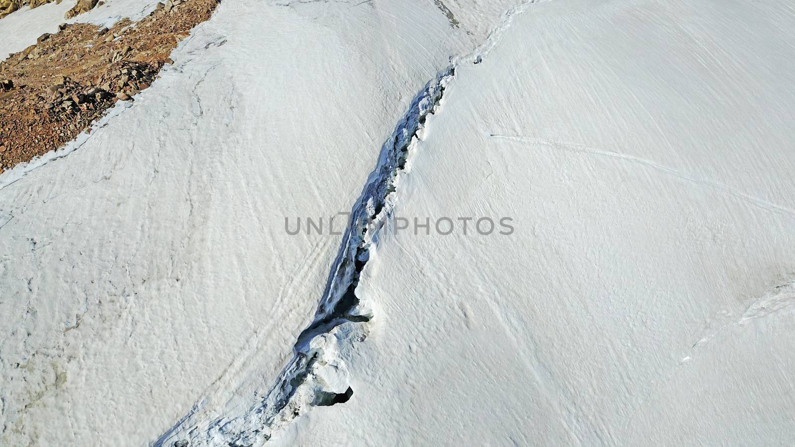 Huge cracks in the glacier at the top of the peak. A fault and a danger of avalanche. You can fall through the ice. Top view from a drone. Mountainous terrain, lots of snow and cliffs. Climbers.
