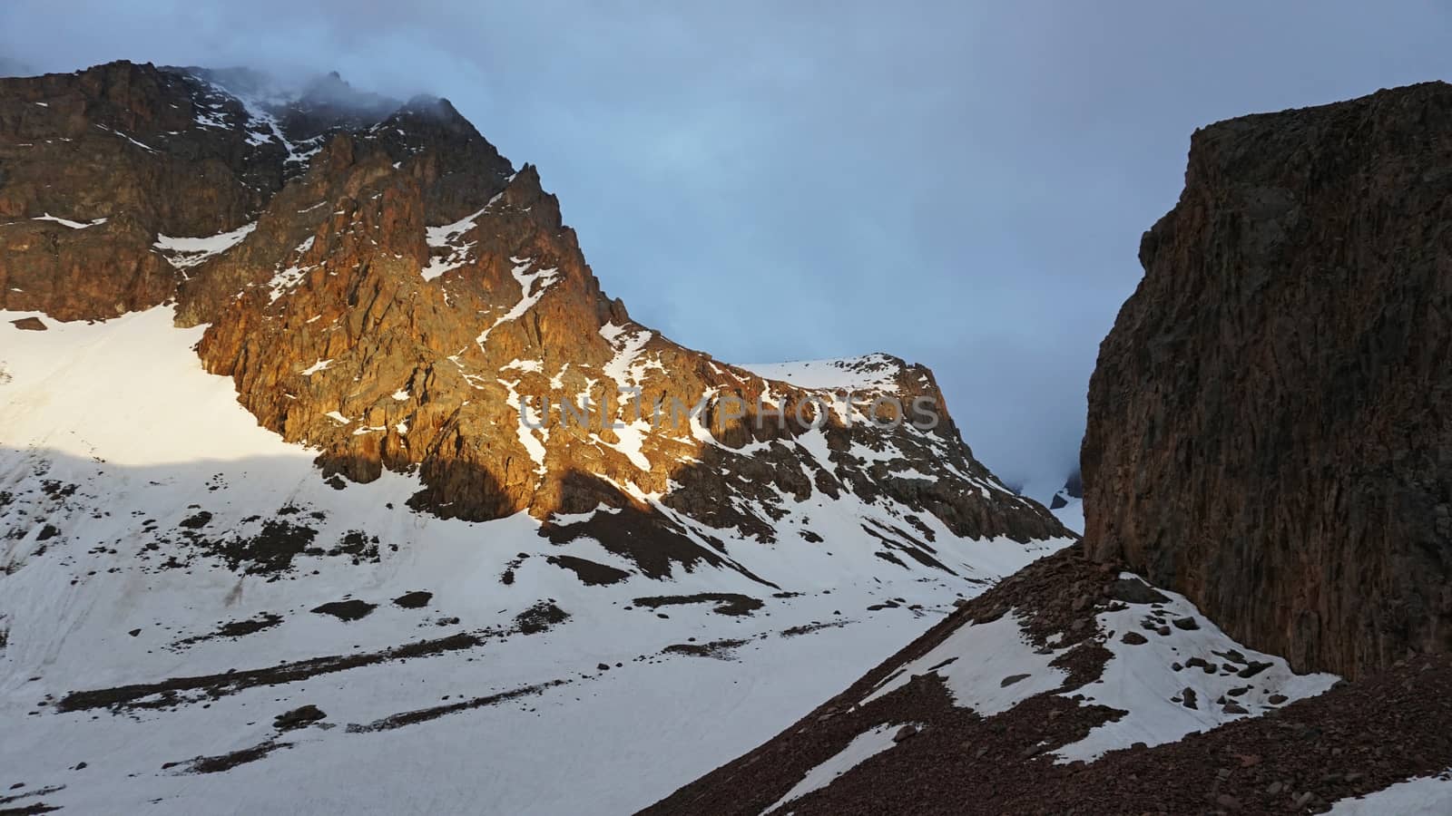 View of the rocky slopes covered with snow. The sun's rays and clouds. Mountaineering camp in the mountains. There is a nunatak. Overnight in the mountains. Sunset in the cold mountains.