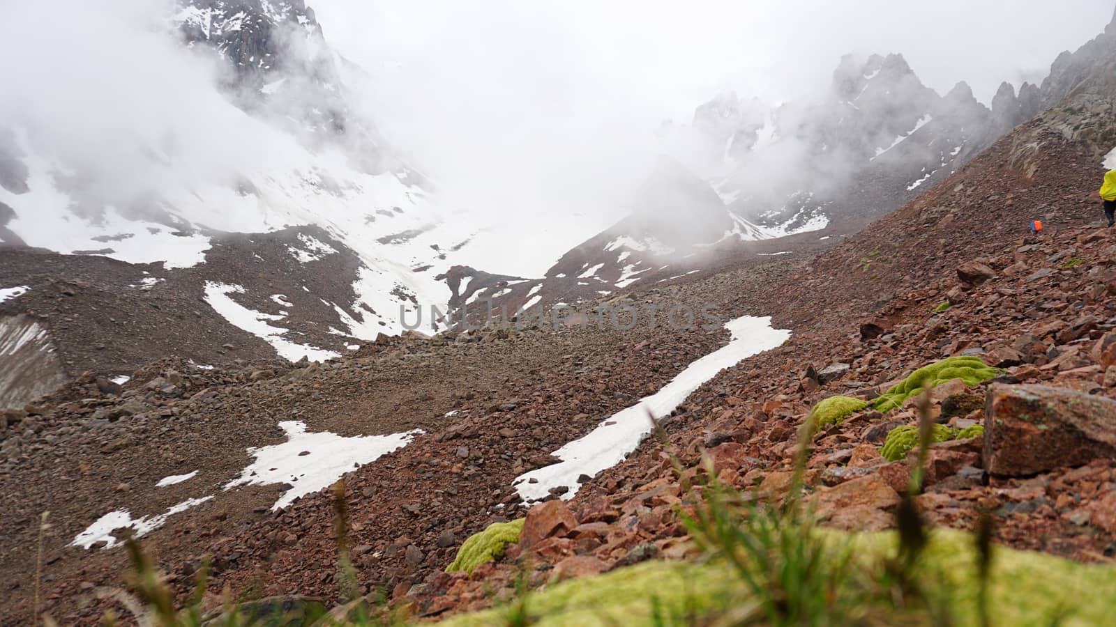 Huge clouds in snowy mountains and green moss. Moss and green grass grow on the rocks. In the fog are climbers. Large rocks and stones. Outdoor activity. Extreme Hiking among clouds and snow peaks.