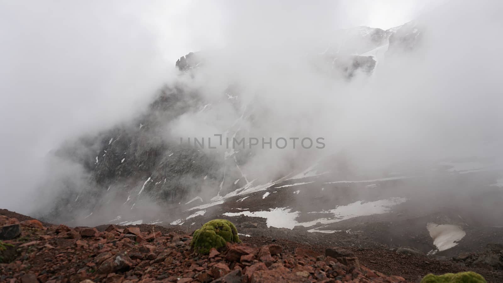 White clouds and snowy peaks. A lot of big stones. Steep cliffs, sometimes covered with white snow. Clouds float through the mountain tops. Moss grows in places. Mountains of the TRANS-ili Alatau.