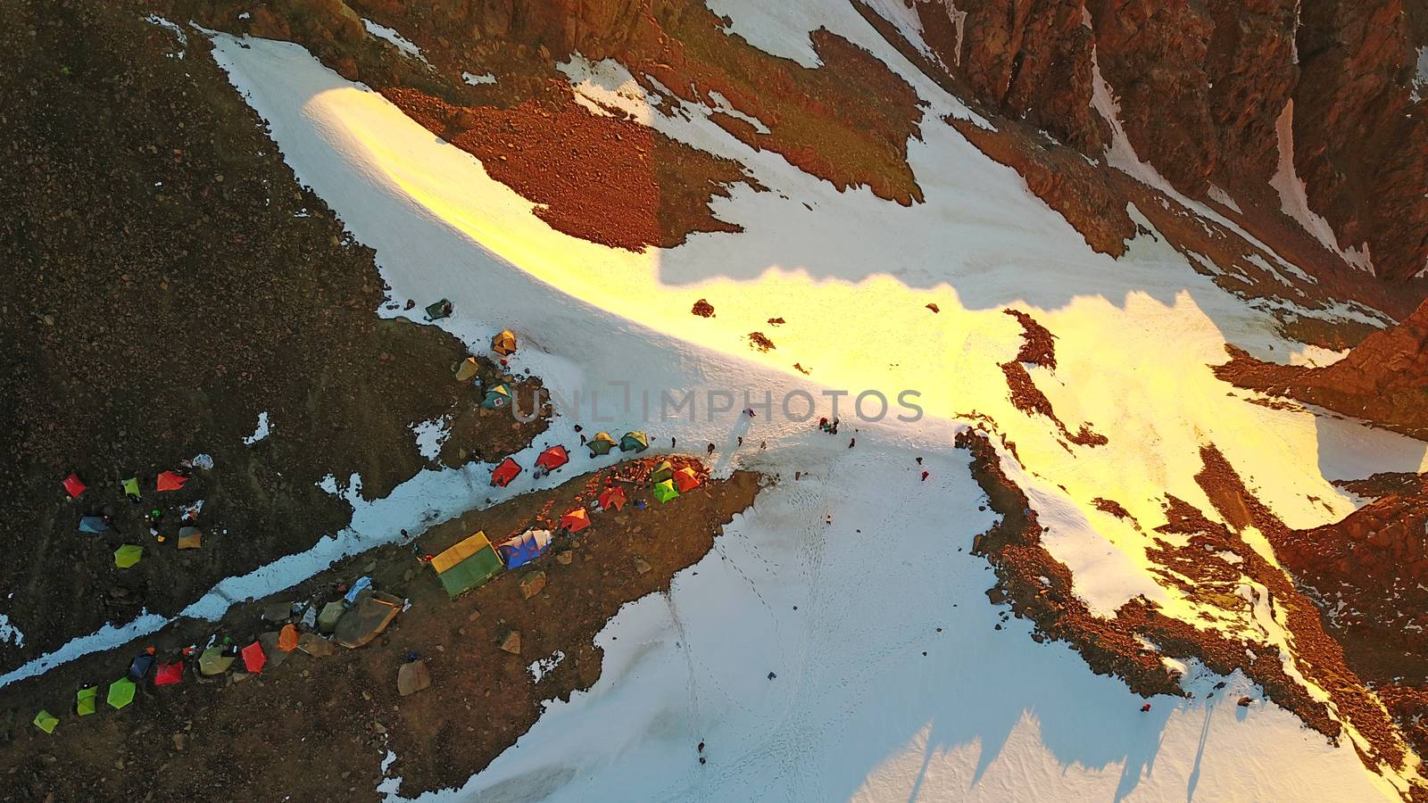 Mountaineering camp high in the snowy mountains. Epic red dawn, top view from a drone. Camp on the edge of the cliff. The snow-capped peaks, huge rocks. Preparing to climb the peak. Lots of tents.