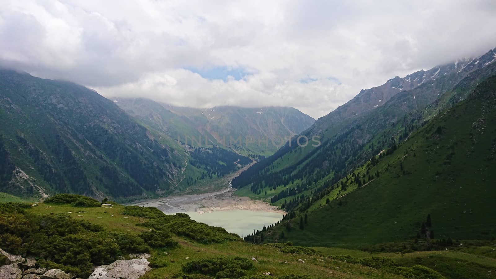 Big Almaty lake in the mountains. Green hills, flowers and fields. White clouds over the mountains. Mountain lake. In places there are stones. Protected place of nature, reserve of TRANS-ili Alatau