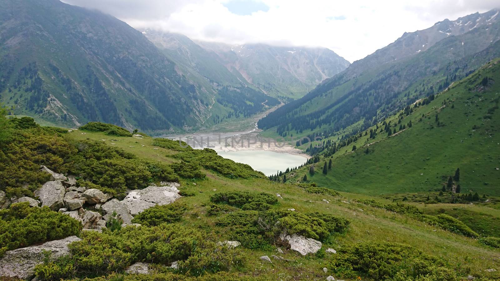 Big Almaty lake in the mountains. Green hills, flowers and fields. White clouds over the mountains. Mountain lake. In places there are stones. Protected place of nature, reserve of TRANS-ili Alatau