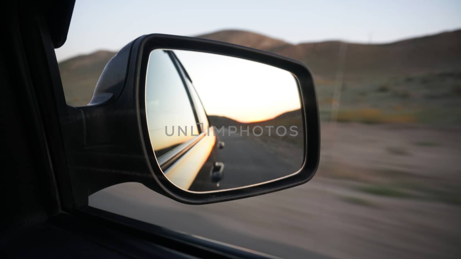 View in the side mirror of the car. Orange dawn over the hills. The car is going at high speed. Green fields, grass, and meadows are visible. Black color of the car.