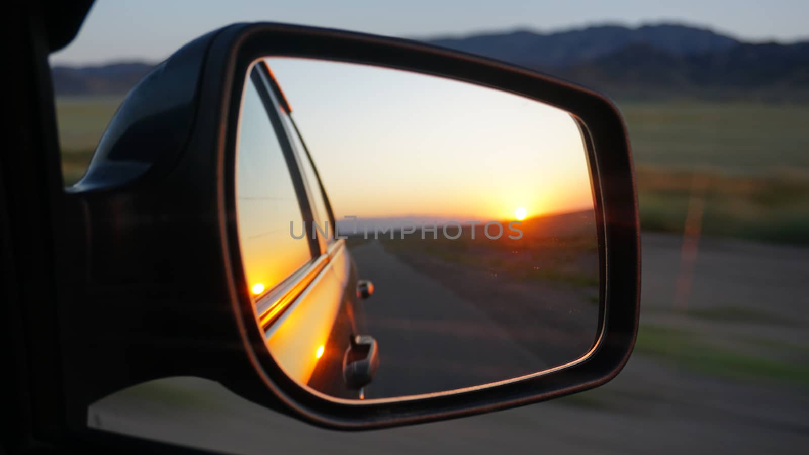 View in the side mirror of the car. Orange dawn over the hills. The car is going at high speed. Green fields, grass, and meadows are visible. Black color of the car.