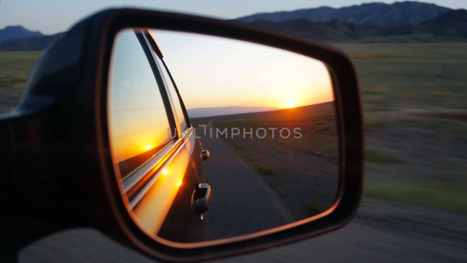 View in the side mirror of the car. Orange dawn over the hills. The car is going at high speed. Green fields, grass, and meadows are visible. Black color of the car.