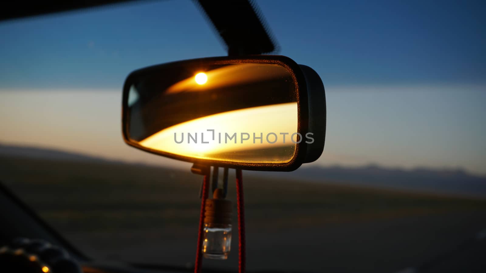 The rear view mirror of the car and dawn. Orange dawn over the hills. The car is going at high speed. Green fields, grass and meadows are visible. Black color of the car. Steering wheel, hood, glass.