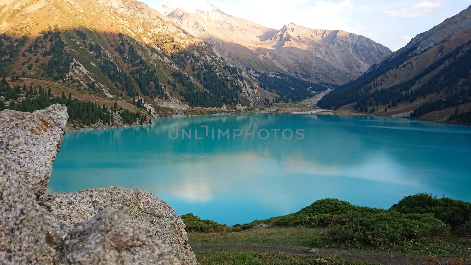 Big Almaty lake in the mountains. Green hills, flowers and fields. White clouds over the mountains. Mountain lake. In places there are stones. Protected place of nature, reserve of TRANS-ili Alatau