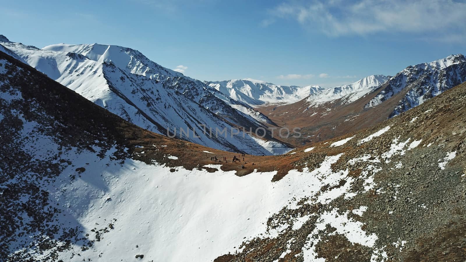 A group of tourists stands on the mountainside. There is snow in places. Very large stones on the sides. Blue sky with white clouds. Top view from the throne. Mountainous area of Kazakhstan.