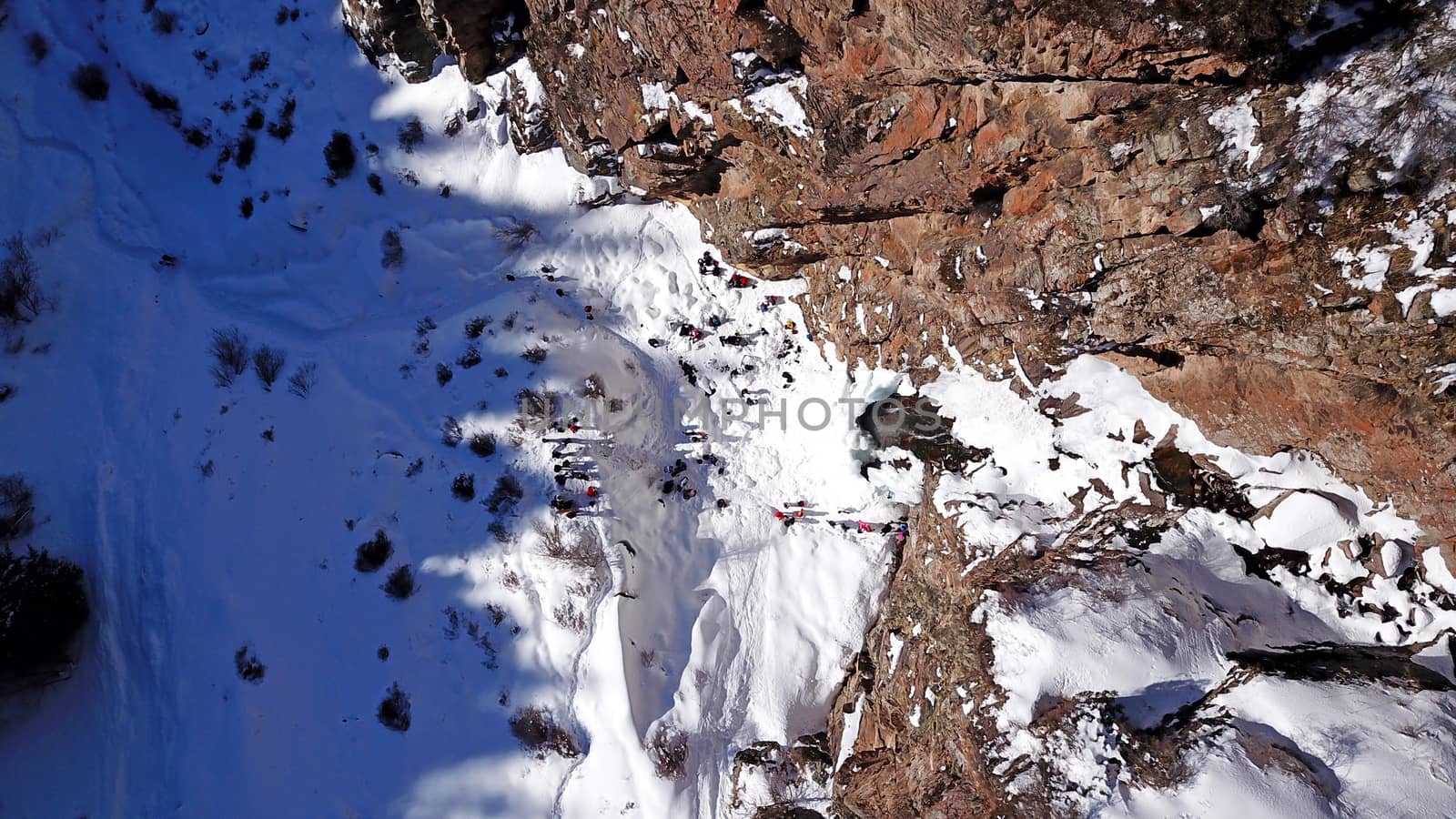 Freezing waterfall in the snowy mountains. View from the drone, from above. The rocks are covered with snow and ice. A small stream of water runs. The waterfall freezes. A group of people are resting