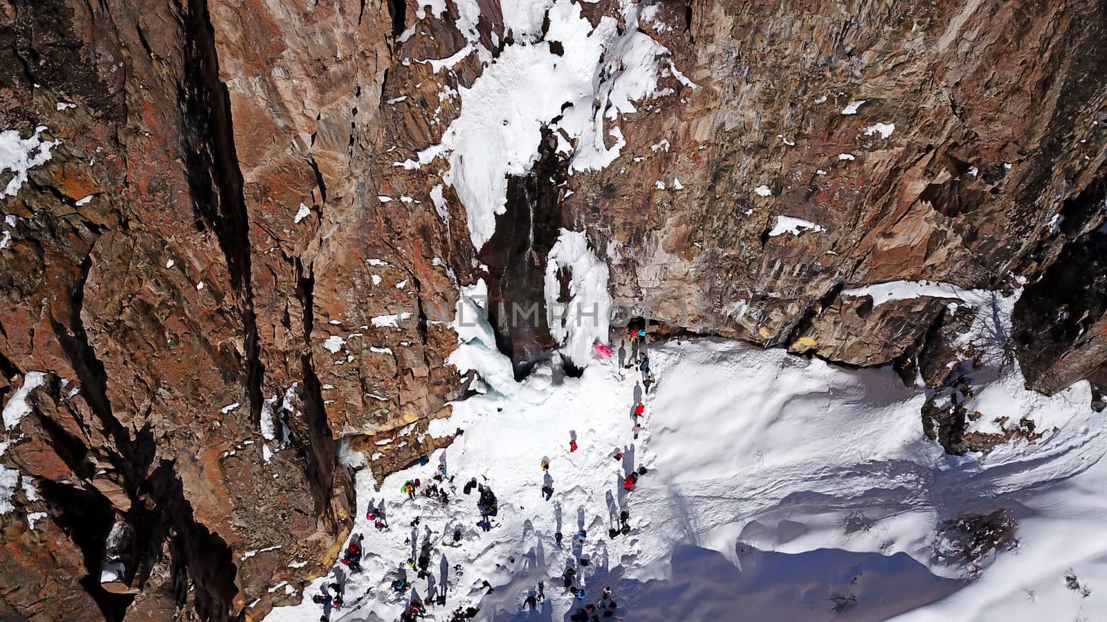 Freezing waterfall in the snowy mountains. View from the drone, from above. The rocks are covered with snow and ice. A small stream of water runs. The waterfall freezes. A group of people are resting