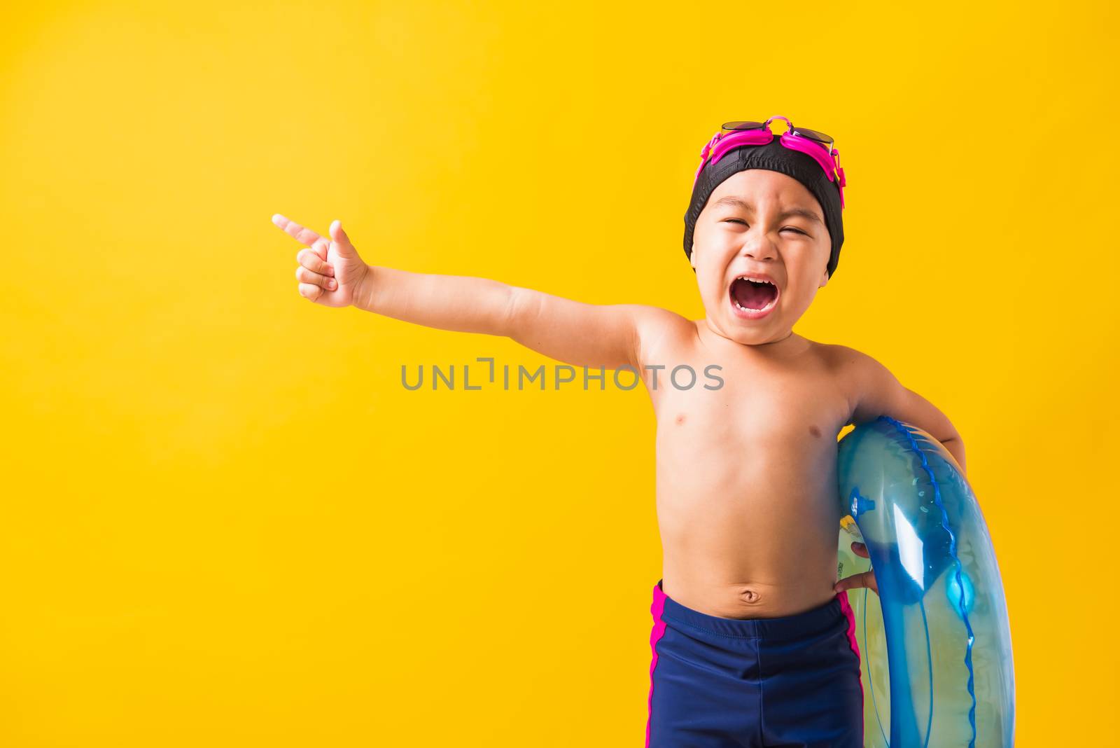 Summer vacation concept, Portrait Asian happy cute little child boy wear goggles and swimsuit hold blue inflatable ring, Kid hav fun point finger to side away, studio shot isolated yellow background