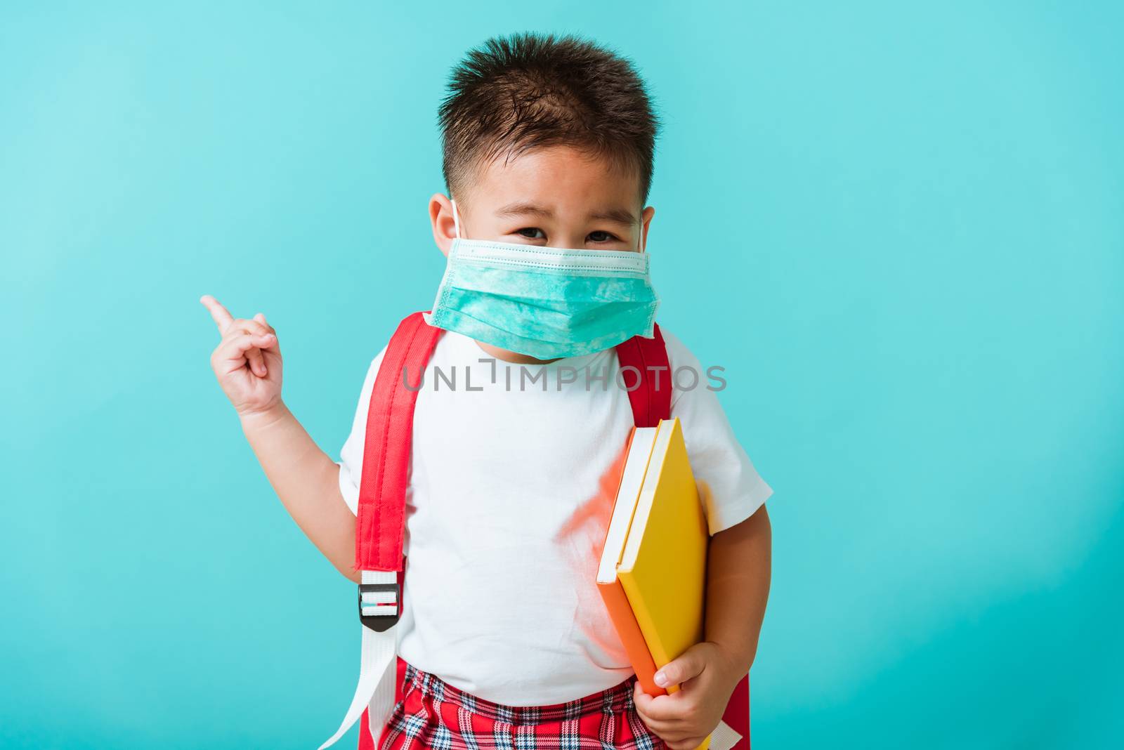 Back to school coronavirus Covid-19 education. Portrait Asian little child boy kindergarten wear face mask protective and school bag hold book before going to school pointing to side away, studio shot