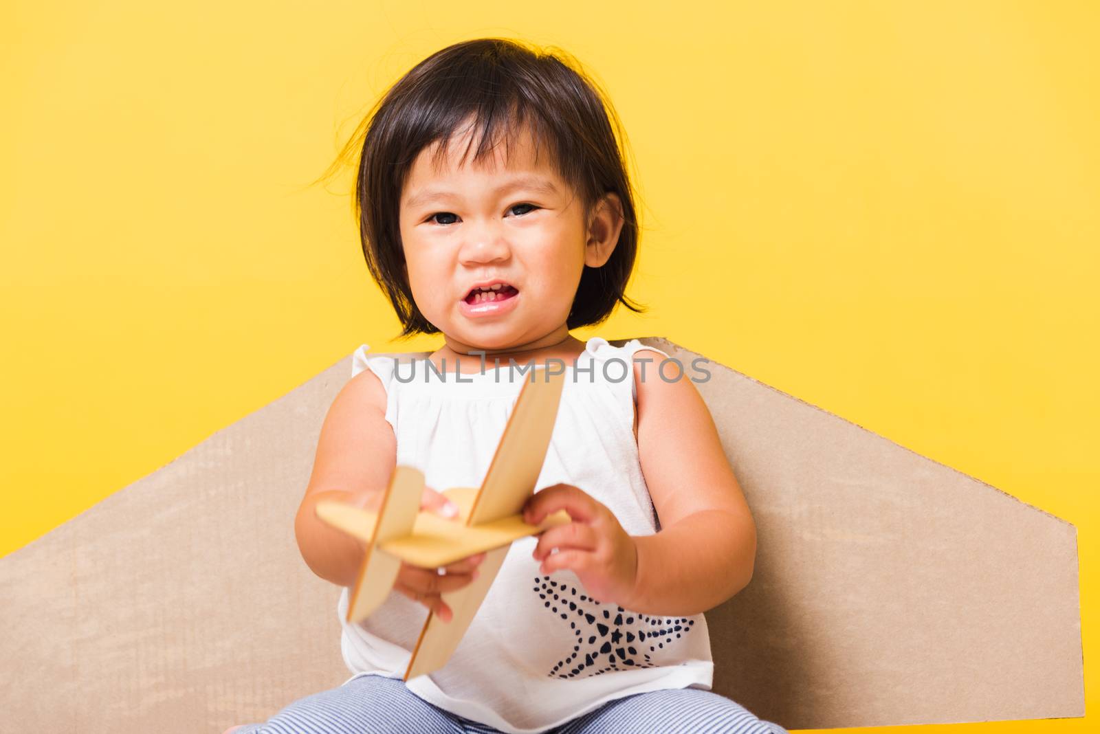 Happy Asian beautiful funny baby little girl smile wear pilot hat play with toy cardboard airplane wings fly hold plane toy, studio shot isolated yellow background, Startup freedom concept