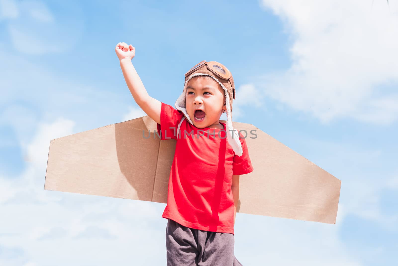 Happy Asian funny child or kid little boy smile wear pilot hat and goggles play toy cardboard airplane wing flying raises hand up against summer blue sky cloud background, Startup freedom concept