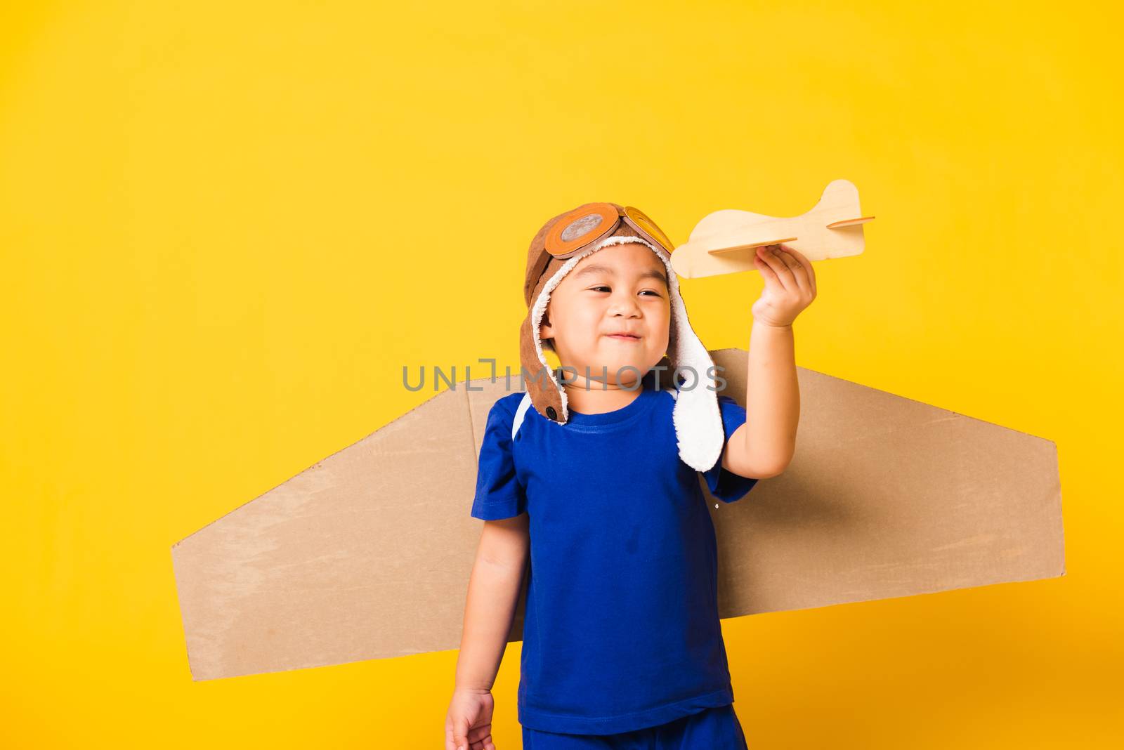 Happy Asian handsome funny child or kid little boy smile wear pilot hat play and goggles with toy cardboard airplane wings fly hold plane toy, studio shot isolated yellow background, Startup freedom