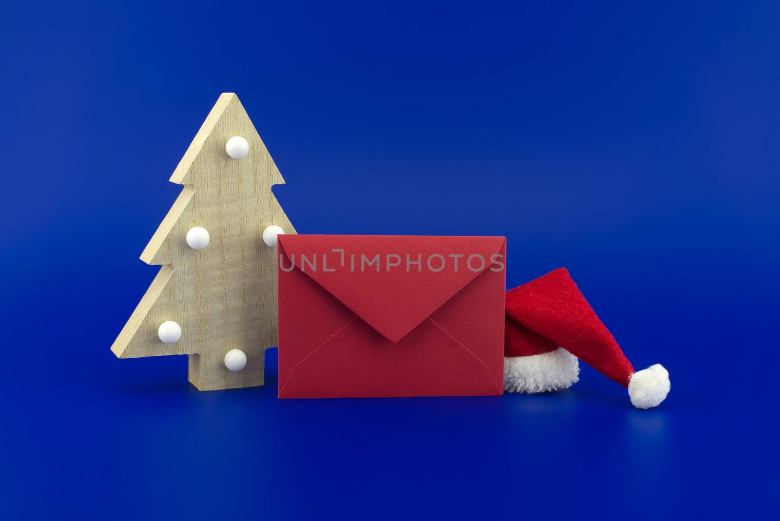 Red envelope for greetings next to a Christmas tree and Santa hat on a festive blue background. New Year and Christmas greeting season concept