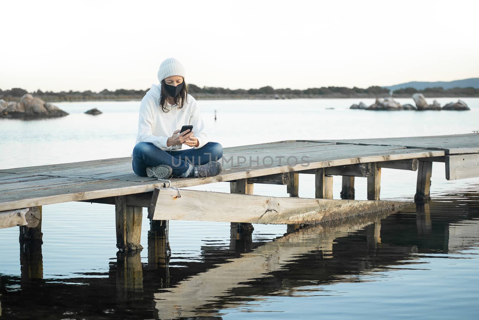 Lonely woman lives nature sitting on a pier at sunset using smartphone to chatting wearing Coronavirus safe protection medical mask - New communication with technology and social network