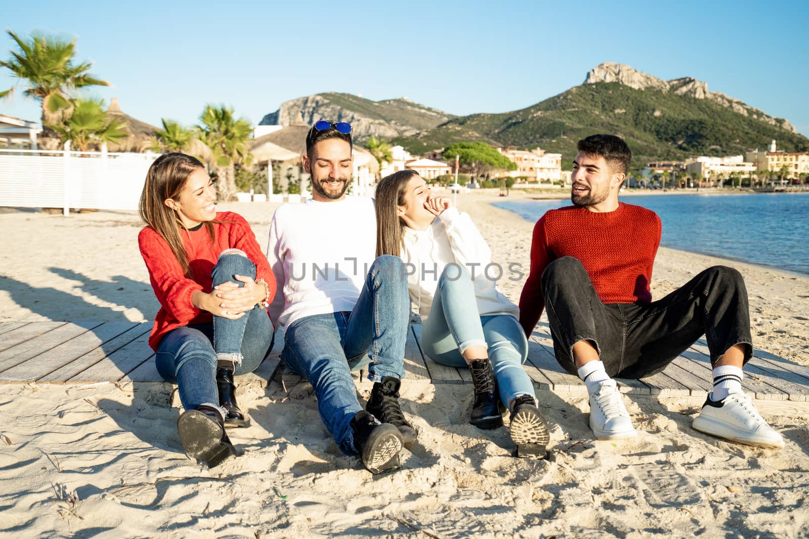 Two young couples by the sea at sunset in a seaside resort in winter having fun talking and laughing each other - Group of friends sitting on the beach in winter sea vacation - White and red clothing by robbyfontanesi