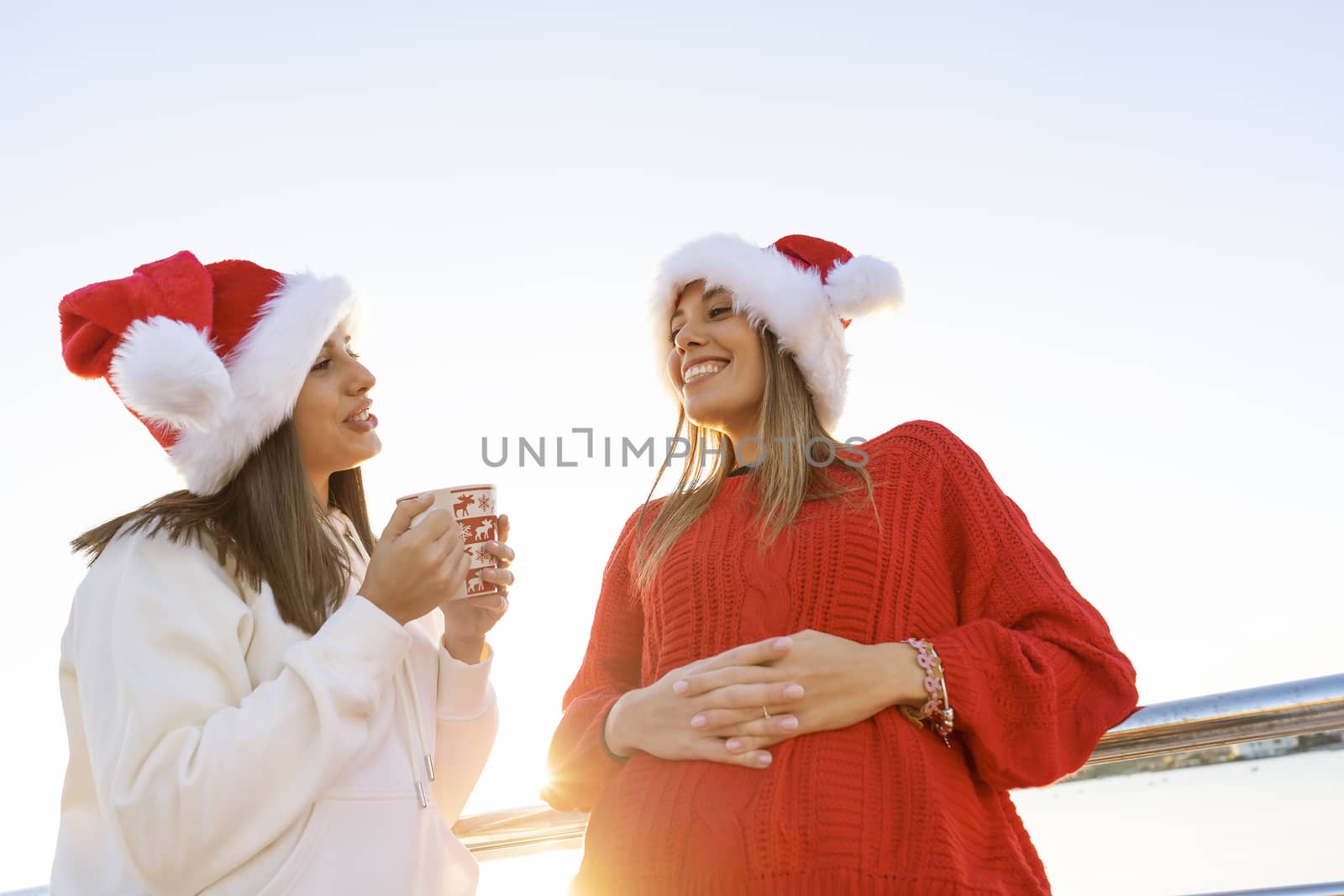Two women in Santa Claus hat having bonding outdoor holding a Christmas tea mug - Bottom view of beautiful woman leaning to a metal railing talking to her best female friend in sunset with reflections by robbyfontanesi