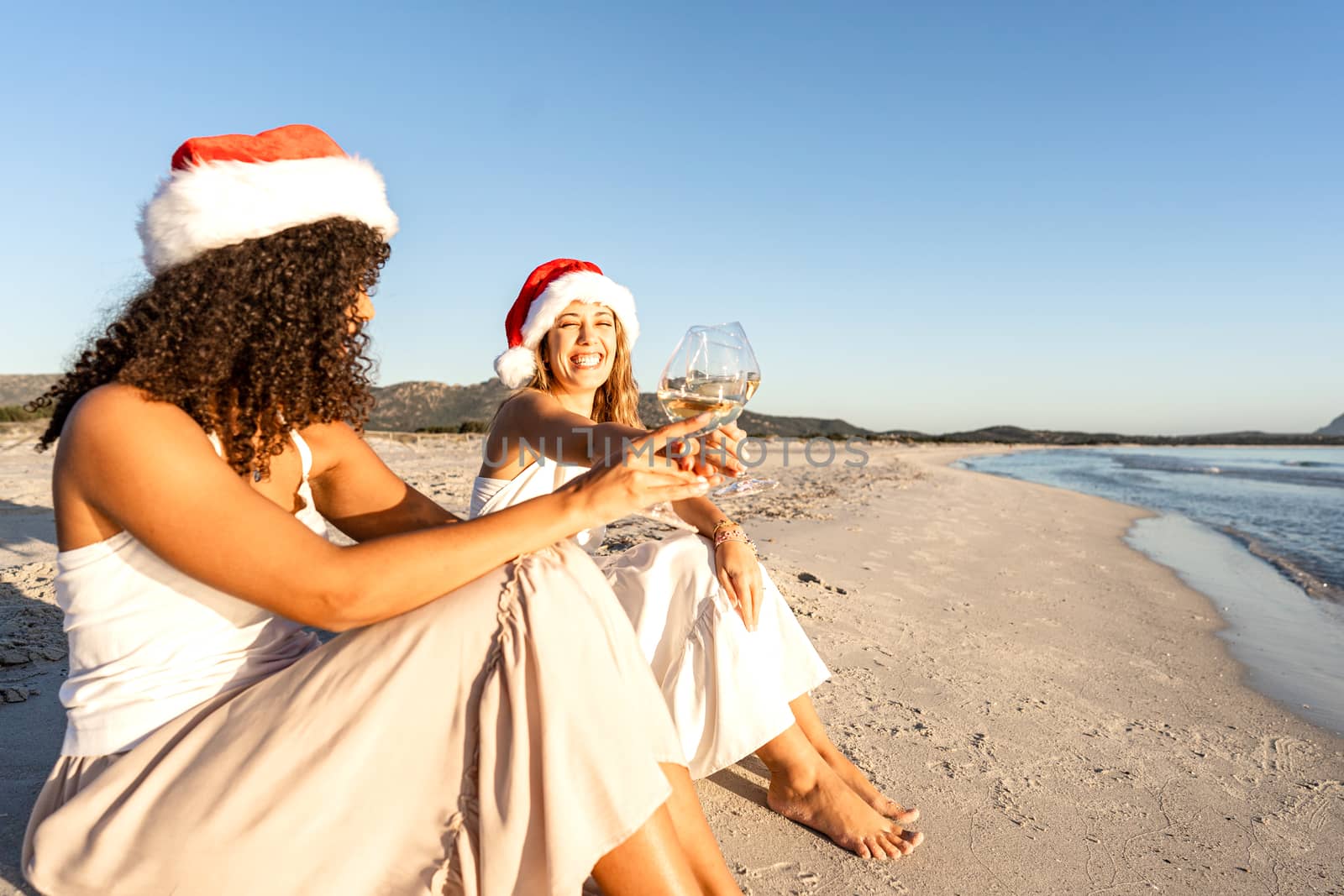 Lesbian couple with Santa hat sitting on seashore toasting with white wine glasses in winter vacation - Two beautiful happy women in love celebrating her Christmas and new year event on the beach by robbyfontanesi