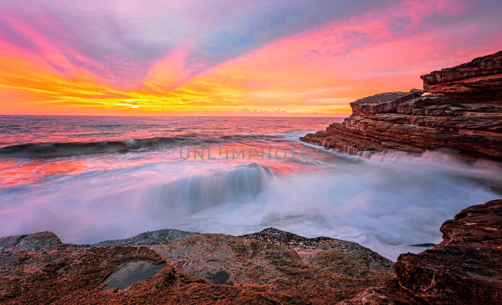 Sunrise sun rays light up the sky and reflect in the ocean and rocks in brilliant vivid colours of orange, yellow and red, while waves crash over large rocks 