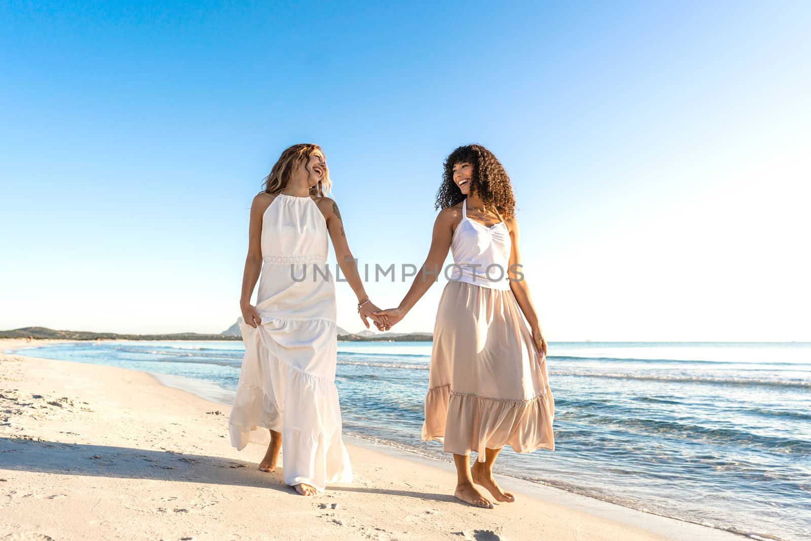 Tattoed beautiful blonde young Caucasian woman laughing holding the hand of her Black Hispanic girlfriend walking in the seashore in the morning or sunset - The beauty of different love between women by robbyfontanesi