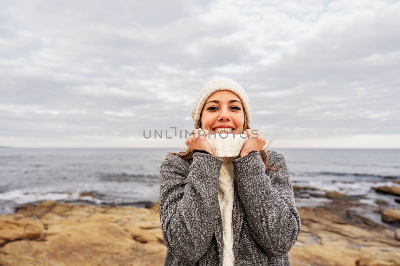 Beautiful young Caucasian woman tightens the neck of her white sweater with her hands for the cold winter weather smiling looking at the camera on sea rocks of Sardinia vacation resort with cloudy sky