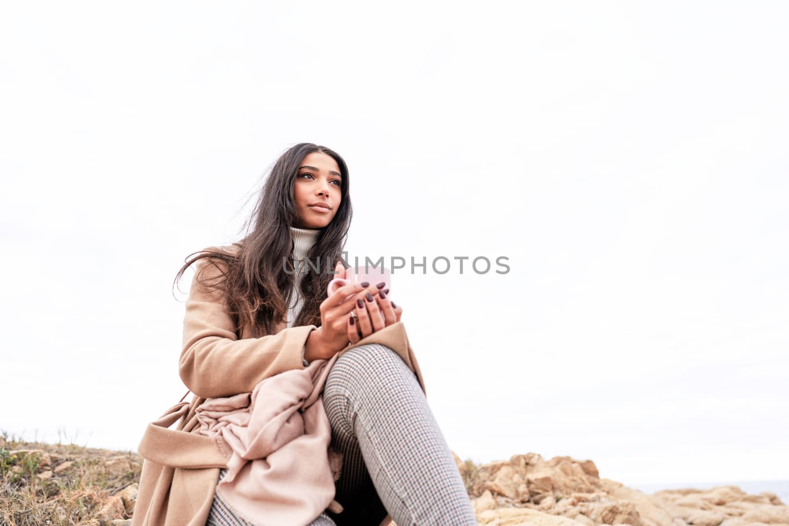 Glamour picture of a beautiful black Hispanic long hair woman sitting on a sea rock holding a mug looking at horizon - Pensive cute stylish female person view from bottom with large white copy space by robbyfontanesi